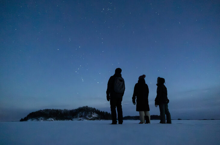 Kolme ihmistä seisovat järven jäällä ja katsovat tähtitaivasta